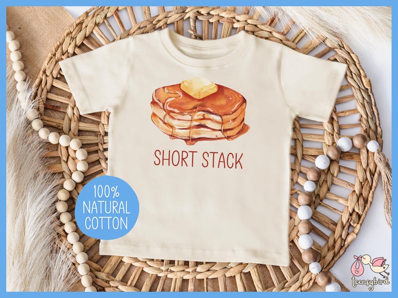 Op de afbeelding: Cr&egrave;mekleurig peuter-T-shirt met een aquarel illustratie van een stapel pannenkoeken met boter en siroop. De tekst "SHORT STACK" staat onder de afbeelding. Een blauwe cirkel met de tekst "100% NATURAL COTTON" staat op het shirt.