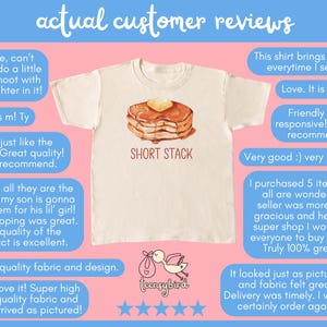 Op de afbeelding: Cr&egrave;mekleurig T-shirt met een illustratie van een stapel pannenkoeken en de tekst "SHORT STACK". De afbeelding is omgeven door klantbeoordelingen. Het T-shirt is een casual kledingstuk.