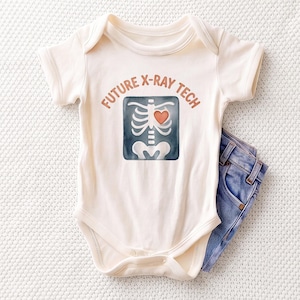 May include: A cream-colored short-sleeve baby bodysuit featuring a graphic of a skeleton rib cage with a red heart and the text FUTURE X-RAY TECH in brown letters, paired with a pair of blue denim jeans tucked underneath the garment.