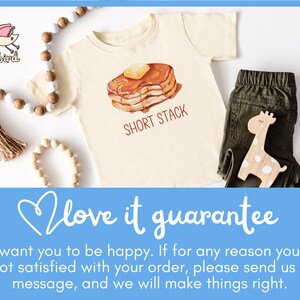 Op de afbeelding: Cr&egrave;mekleurig peuter-T-shirt met een pannenkoekenafbeelding en de tekst "SHORT STACK". Het shirt wordt gecombineerd met een olijfgroene broek en een houten girafespeelgoed. Decoratieve kralen en een kwast zijn ook aanwezig.