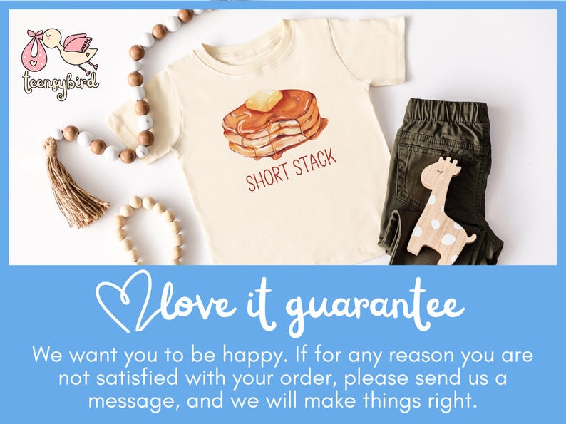 Op de afbeelding: Cr&egrave;mekleurig peuter-T-shirt met een pannenkoekenafbeelding en de tekst "SHORT STACK". Het shirt wordt gecombineerd met een olijfgroene broek en een houten girafespeelgoed. Decoratieve kralen en een kwast zijn ook aanwezig.