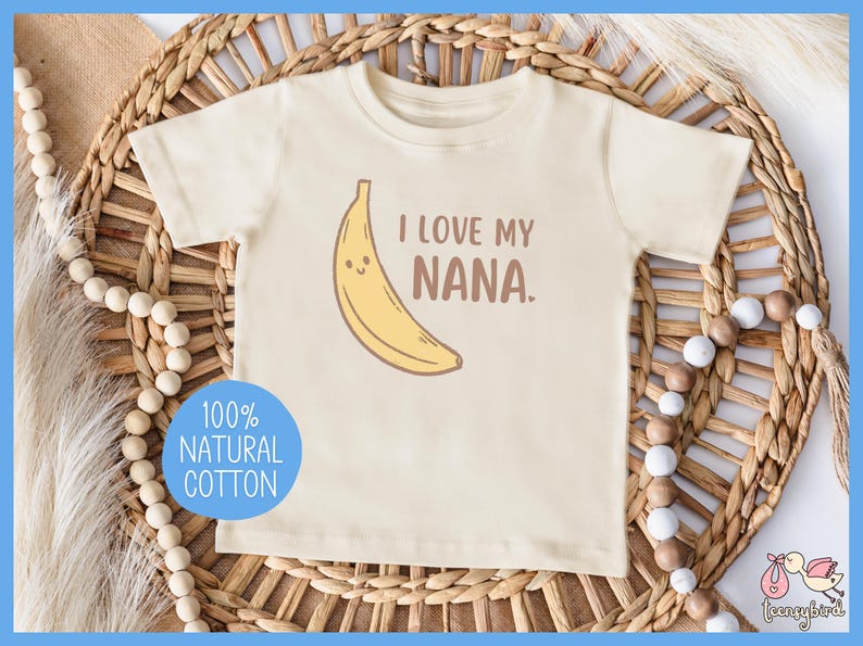 May include: Cream-colored toddler t-shirt with a smiling banana graphic and the text "I LOVE MY NANA." A blue circle with the words "100% NATURAL COTTON" is on the bottom left. The shirt is on a woven background.