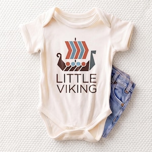 May include: A cream-colored short-sleeve baby bodysuit featuring a graphic of a stylized Viking ship with a red and blue sail. Below the ship, the text reads LITTLE VIKING in dark brown capital letters. A pair of blue denim jeans is tucked underneath.
