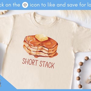 Op de afbeelding: Cr&egrave;mekleurig T-shirt met een aquarel illustratie van een stapel pannenkoeken met boter en siroop. De tekst "SHORT STACK" is in het rood gedrukt. Het shirt maakt deel uit van een kledingcollectie.