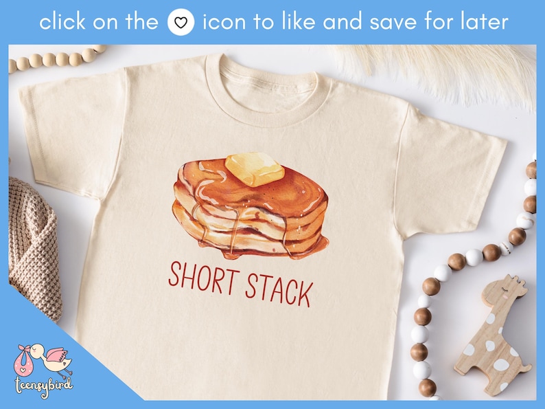 Op de afbeelding: Cr&egrave;mekleurig T-shirt met een aquarel illustratie van een stapel pannenkoeken met boter en siroop. De tekst "SHORT STACK" is in het rood gedrukt. Het shirt maakt deel uit van een kledingcollectie.