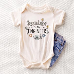 May include: A cream-colored short-sleeve baby bodysuit featuring the text Assistant to the ENGINEER in black, accompanied by illustrations of two yellow light bulbs, three colorful gears in gray and orange, and a small gray robot holding a wrench.