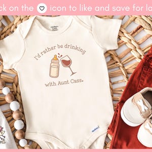 May include: Cream-colored baby onesie with the text "I'd rather be drinking with Aunt Cass." A bottle and wine glass graphic are printed on the front. Paired with rust-colored pants and baby shoes.