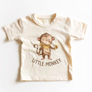 May include: A cream-colored short-sleeve t-shirt featuring a brown watercolor illustration of a monkey wearing a yellow neckerchief and holding a yellow banana. Below the monkey, the text LITTLE MONKEY is printed in brown capital letters.