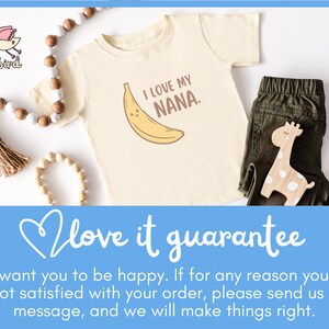 May include: Cream-colored baby tee with a banana graphic and the text "I LOVE MY NANA." The tee is paired with olive green pants and a wooden giraffe toy. A beaded garland and a tassel are also in the image.