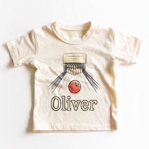May include: A cream-colored short-sleeve t-shirt featuring a graphic illustration of a bowling lane with pins and a red bowling ball. The name Oliver is printed in a bold, dark font below the bowling graphic on the front of the garment.