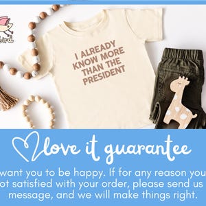 May include: A light beige baby onesie with the text "I ALREADY KNOW MORE THAN THE PRESIDENT" printed in brown. The onesie is lying on a white surface with wooden beads and a wooden giraffe toy.