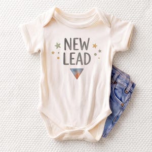 May include: A cream-colored short-sleeve baby bodysuit featuring the text NEW LEAD in dark gray letters, accented by small green and yellow stars and dots, with a single downward-pointing triangle graphic below the text and blue denim jeans.