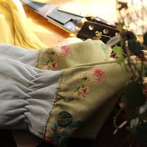 May include: A pair of light gray leather gloves with a floral patterned cuff. The cuff features a light green fabric with pink and red roses. A yellow garment and leather tools are visible in the background.