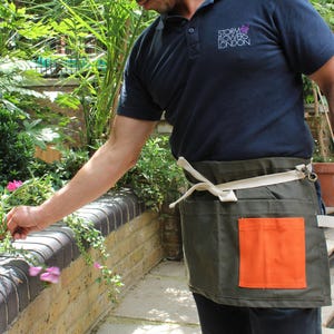 May include: A dark olive green waist apron featuring a bright orange rectangular pocket on the front and a cream-colored adjustable waist tie with a metal hook closure. The apron is worn over a dark navy blue polo shirt with white text.