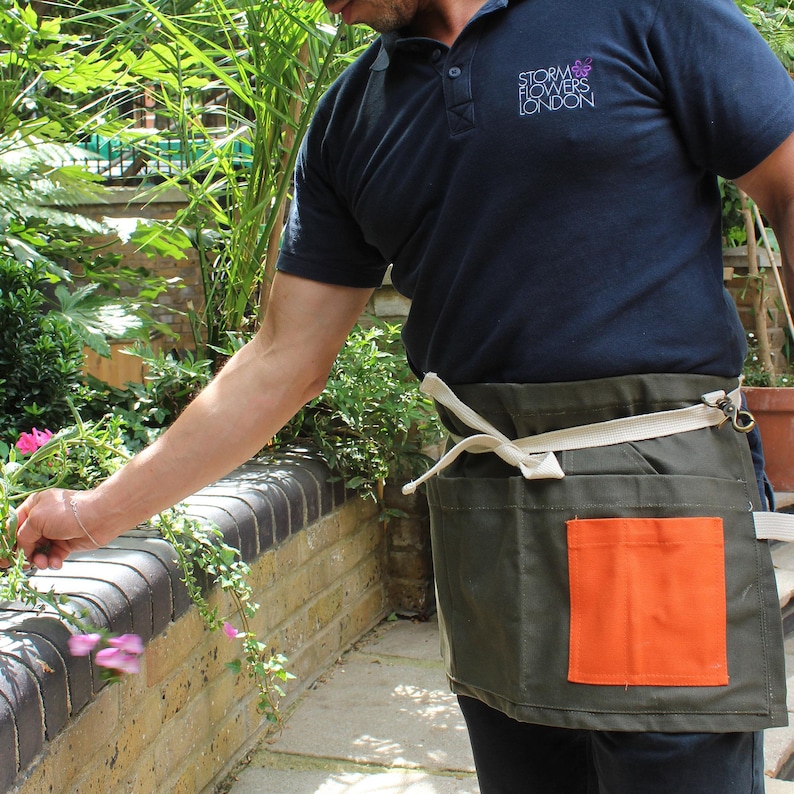 May include: A dark olive green waist apron featuring a bright orange rectangular pocket on the front and a cream-colored adjustable waist tie with a metal hook closure. The apron is worn over a dark navy blue polo shirt with white text.