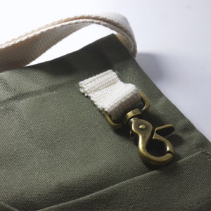 May include: An olive green canvas fabric bag featuring a thick, off-white woven cotton strap attached to a metallic brass-toned swivel lobster clasp. The hardware is secured to the bag with a matching off-white fabric loop stitched onto the surface.