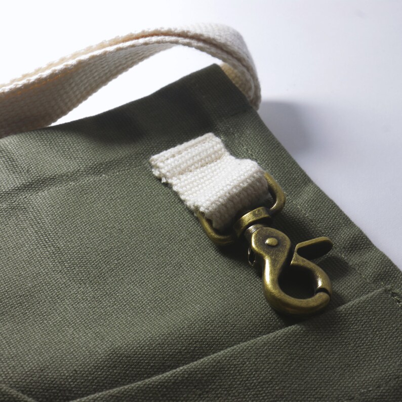 May include: An olive green canvas fabric bag featuring a thick, off-white woven cotton strap attached to a metallic brass-toned swivel lobster clasp. The hardware is secured to the bag with a matching off-white fabric loop stitched onto the surface.