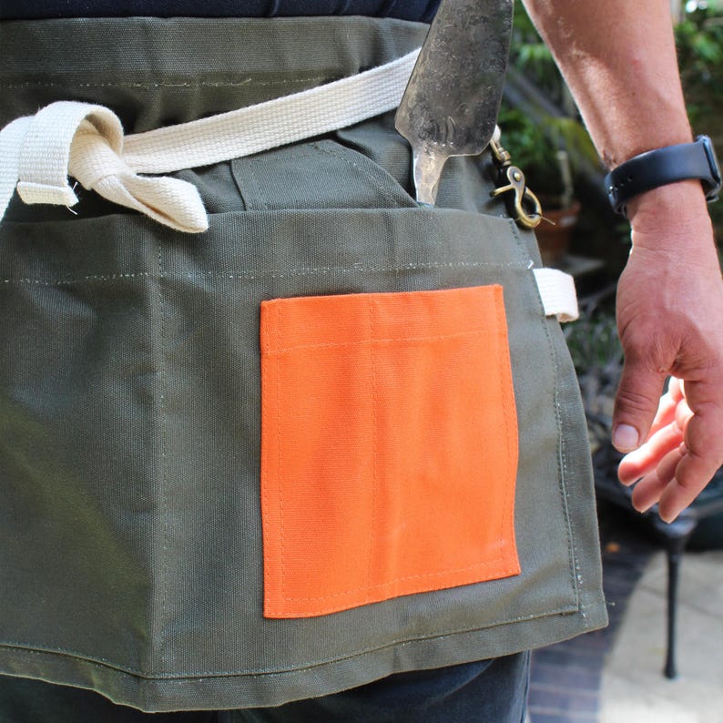 May include: An olive green utility apron featuring a bright orange rectangular pocket on the front, multiple storage compartments, and a thick off-white woven waist tie. A metal garden trowel is tucked into one of the upper pockets.