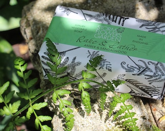 May include: A white bar of soap with a green and white label that says "Storm Flowers Garden Rake & Lather Amber & Sandalwood" and "Luxury Vegetable Soap Made in England". The soap is on a gray stone with green ferns.