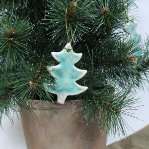 Puede incluir: Un adorno de árbol de Navidad de cerámica azul con detalles de purpurina dorada colgado de un árbol de Navidad artificial verde.