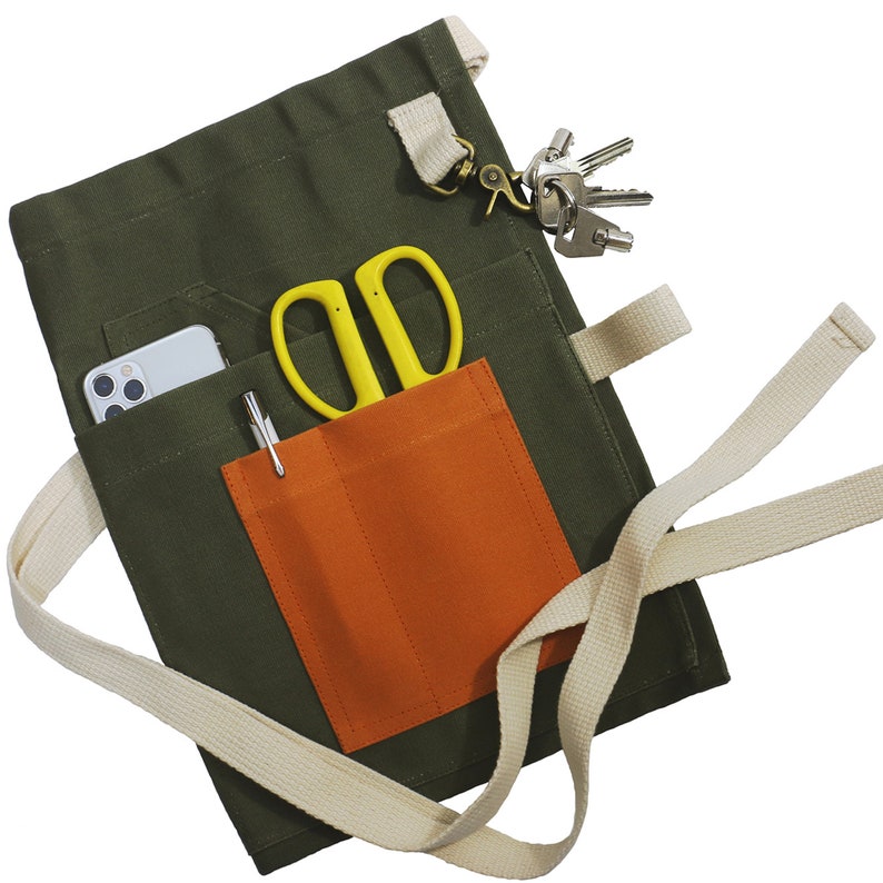 May include: An olive green utility apron featuring a large orange front pocket, a beige waist tie, and a metal key clip. The apron contains a silver smartphone, a pair of bright yellow scissors, and a silver pen tucked into its various pockets.