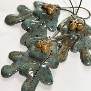 May include: Three ceramic oak leaf ornaments with a glossy teal and blue glaze. Each leaf features two small, textured brown acorns attached near the top stem area. A thin, grey braided cord is looped through a hole at the top of each leaf.