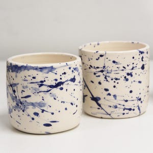 Op de afbeelding: Twee cilindrische keramische bekers met een gebroken witte basiskleur en een donkerblauw verfspatpatroon. De vaten hebben gladde, afgeronde randen en rechte zijkanten, wat een handmatig aangebrachte esthetiek met variërende druppelgroottes toont.