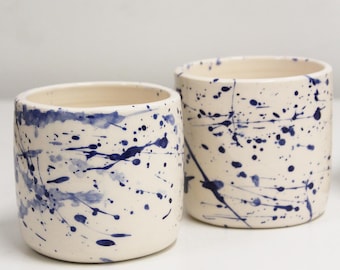 May include: Two cylindrical ceramic cups featuring an off-white base color with a dark blue paint splatter pattern. The vessels have smooth, rounded rims and straight sides, showcasing a hand-applied aesthetic with varying droplet sizes.