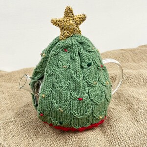 May include: A green knitted Christmas tree tea cozy with a gold star on top. The cozy is decorated with small red and gold accents. The bottom edge is trimmed with red. The cozy is covering a clear glass teapot.