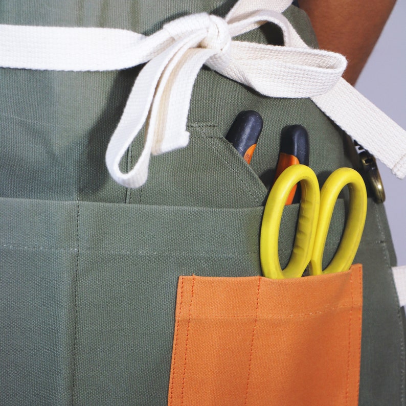 May include: An olive green utility apron featuring a white fabric waist tie, a large rectangular orange front pocket, and additional side pockets. Bright yellow handled scissors are tucked into the orange pocket, with black and orange tools visible.