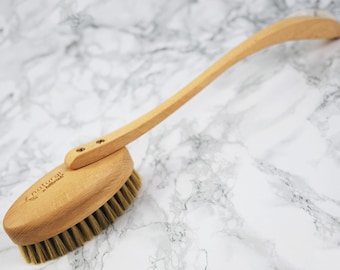 May include: A wooden back brush with a long, curved handle and oval brush head. The brush head features natural bristles and is attached to the handle with two screws. The brush is made of light-colored wood and is designed for body care.