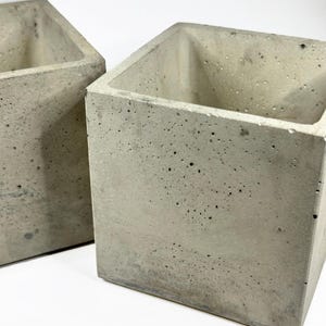Puede incluir: Dos maceteros cuadrados de hormigón gris claro. Los maceteros tienen una superficie rugosa y texturizada con pequeños agujeros y un diseño minimalista. Adecuados para uso en interiores o exteriores.