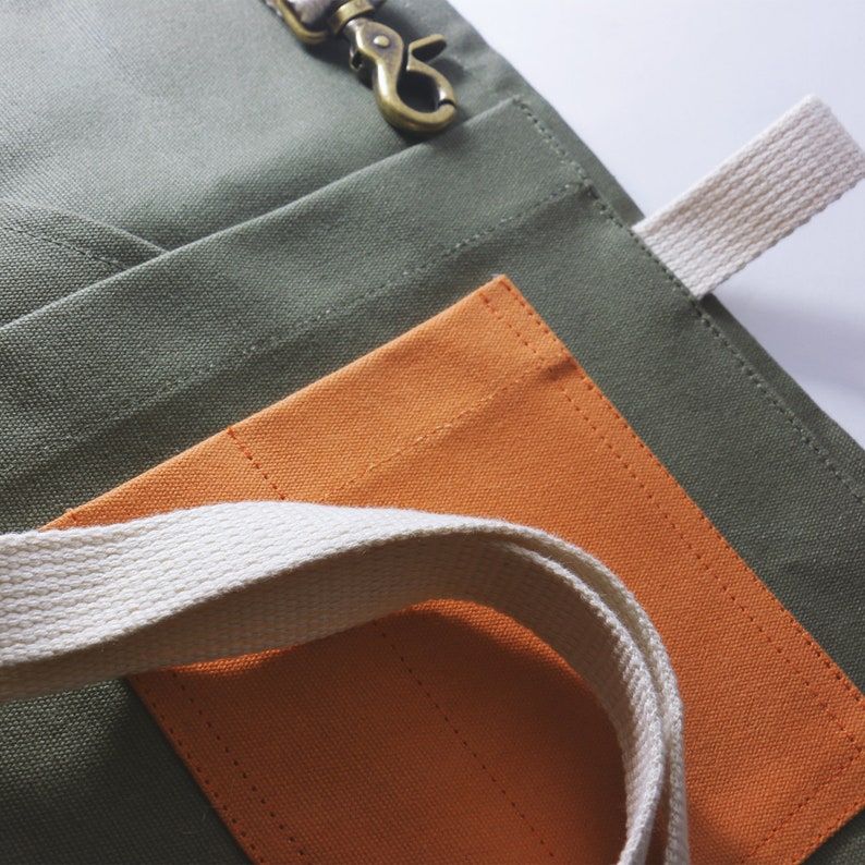 May include: An olive green canvas apron featuring a bright orange rectangular pocket and a cream-colored woven strap. A metallic brass-toned swivel lobster clasp is attached to the top edge of the apron, providing a functional hardware detail.