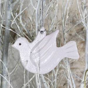 May include: A white ceramic dove ornament with a silver string for hanging. The dove has a textured surface and a slightly iridescent finish.