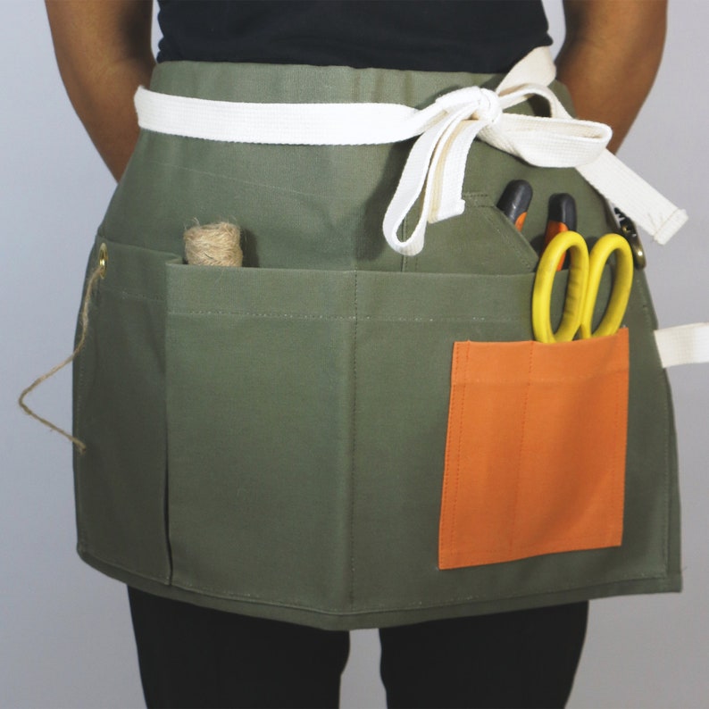 May include: An olive green waist apron featuring a white fabric tie at the waist, multiple storage pockets, and a contrasting orange pocket. The apron contains a roll of twine, a pair of yellow-handled scissors, and other small tools.