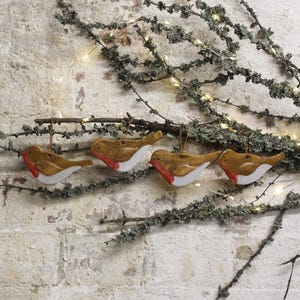 May include: Four ceramic bird ornaments with brown, white, and red details hang from a small branch. The ornaments are suspended against a backdrop of branches with fairy lights and a textured brick wall.