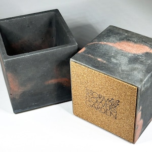 May include: Two square concrete planters. One is open, the other is closed with a cork base. The planters are dark grey with reddish-brown marbling. The cork base has the text "Storm Flowers Garden".