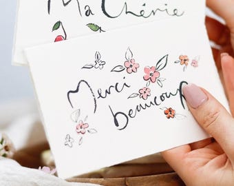 May include: A white card with the words "Merci beaucoup" written in black cursive script. The card is decorated with small pink and orange flowers and green leaves.