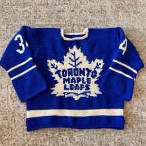 May include: A blue and white crochet hockey jersey with the Toronto Maple Leafs logo. The jersey has white stripes on the sleeves and hem, with the number 34 on the sleeves. The jersey is made of a soft, textured yarn.