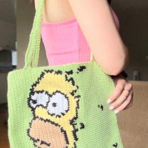 May include: A green crocheted tote bag with a yellow cartoon character on the front. The character has black eyes, a black outline, and a yellow body. The bag has a long green strap.