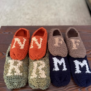 May include: Four pairs of handmade crochet slippers in various colours. One pair is orange with the letter N, one pair is olive green with the letter R, one pair is light brown with the letter F, and one pair is navy blue with the letter M.