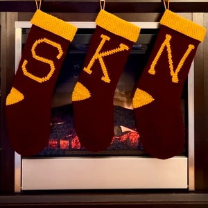 Peut inclure: Trois chaussettes de Noël en crochet rouge et or avec les lettres S, K et N dessus, accrochées au-dessus d'une cheminée.