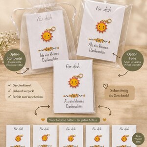 Könnte beinhalten: Eine Sammlung von sonnenförmigen Metallanhängern mit goldenen Karabinerverschlüssen, befestigt auf weißen Karten mit Blumengrafiken und deutschem Text wie "Für dich" und "Als ein kleines Dankeschön," verpackt in transparenten Beuteln.