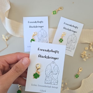 Könnte beinhalten: Eine Hand hält eine Karte mit dem Text "Freundschafts Glücksbringer" und einer Zeichnung von zwei Personen. Ein goldfarbener Anhänger mit einem grünen vierblättrigen Kleeblatt ist an der Karte befestigt. Weitere Karten und Anhänger sind sichtbar.