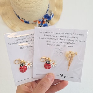 Könnte beinhalten: Zwei Marienkäfer-Anhänger mit Gänseblümchen-Akzenten und goldfarbenen Verschlüssen werden gezeigt. Jeder Anhänger ist an einer Karte mit deutschem Text befestigt. Ein Strohhut mit blauem und weißem Band ist im Hintergrund.
