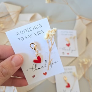 Könnte beinhalten: Ein kleiner roter Herzanhänger mit goldenem Verschluss hängt an einer weißen Karte. Auf der Karte steht in schwarzer Schrift "A LITTLE HUG TO SAY A BIG thank you". Trockenblumen und weitere Karten sind im Hintergrund.