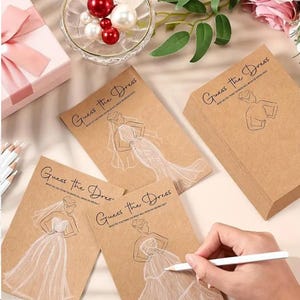 Könnte beinhalten: Ein Set aus braunen Kraftpapierkarten mit dem Text Guess the Dress und einer weißen Strichzeichnung eines Brautkleides. Die Karten sind mit einem Stapel, einer Glasschale mit roten und weißen Perlen und einer rosa Geschenkbox angeordnet.