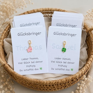 Könnte beinhalten: Zwei weiße Karten mit dem Text 'Glücksbringer', 'Thomas' oder 'Samuel' und 'Lieber Thomas/Samuel, Viel Glück bei deiner Prüfung Du schaffst das'. Jede Karte enthält einen kleinen goldfarbenen Anhänger, ein rosa Schweinchen und ein grünes Kleeblatt.