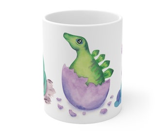 Op de afbeelding: Een witte keramische mok met een groene dinosaurus die uit een paars ei komt. De dinosaurus heeft een groen lichaam, een paarse schil en een groene staart. Er liggen kleine stukjes gebroken schil rond het ei.