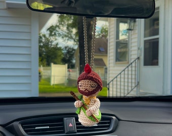 May include: A handmade crochet car charm featuring a figure with a brown hat, beard, and a green and beige outfit. The charm hangs from a silver chain and is suspended in front of a car's dashboard and rearview mirror.